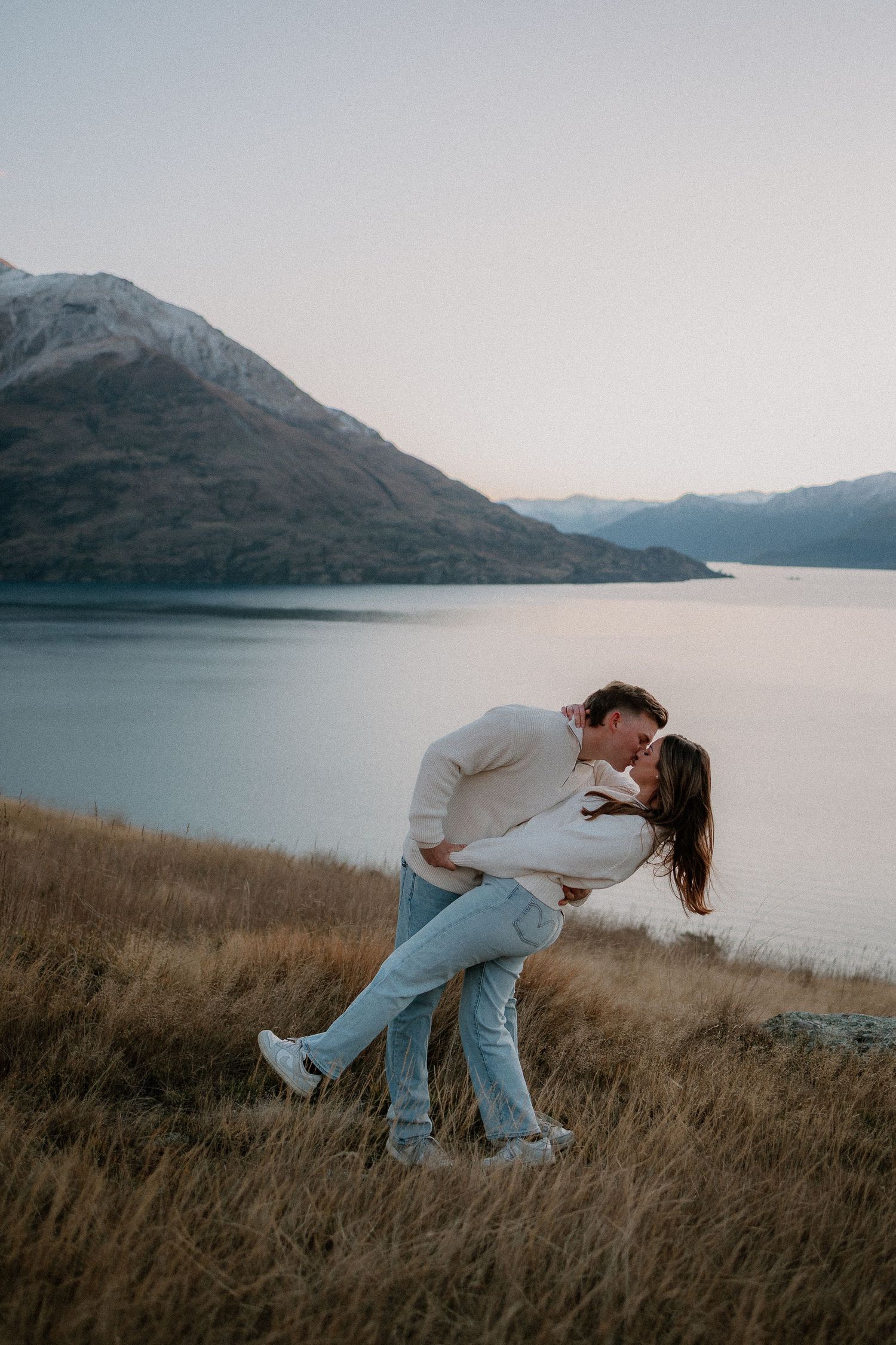 Couple sharing a romantic dip kiss on a hillside overlooking a serene lake and snow-capped mountains at dusk.