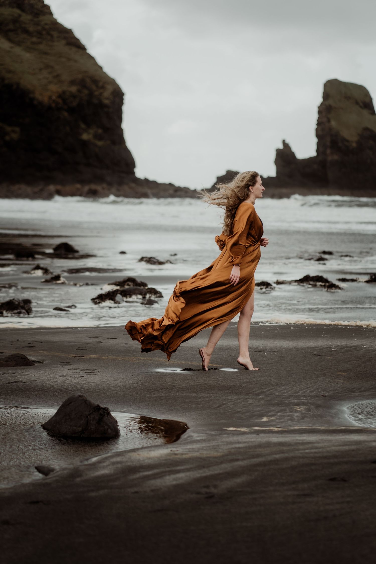 Woman in flowing amber dress walks barefoot on dark volcanic beach with dramatic rock formations and moody ocean waves.