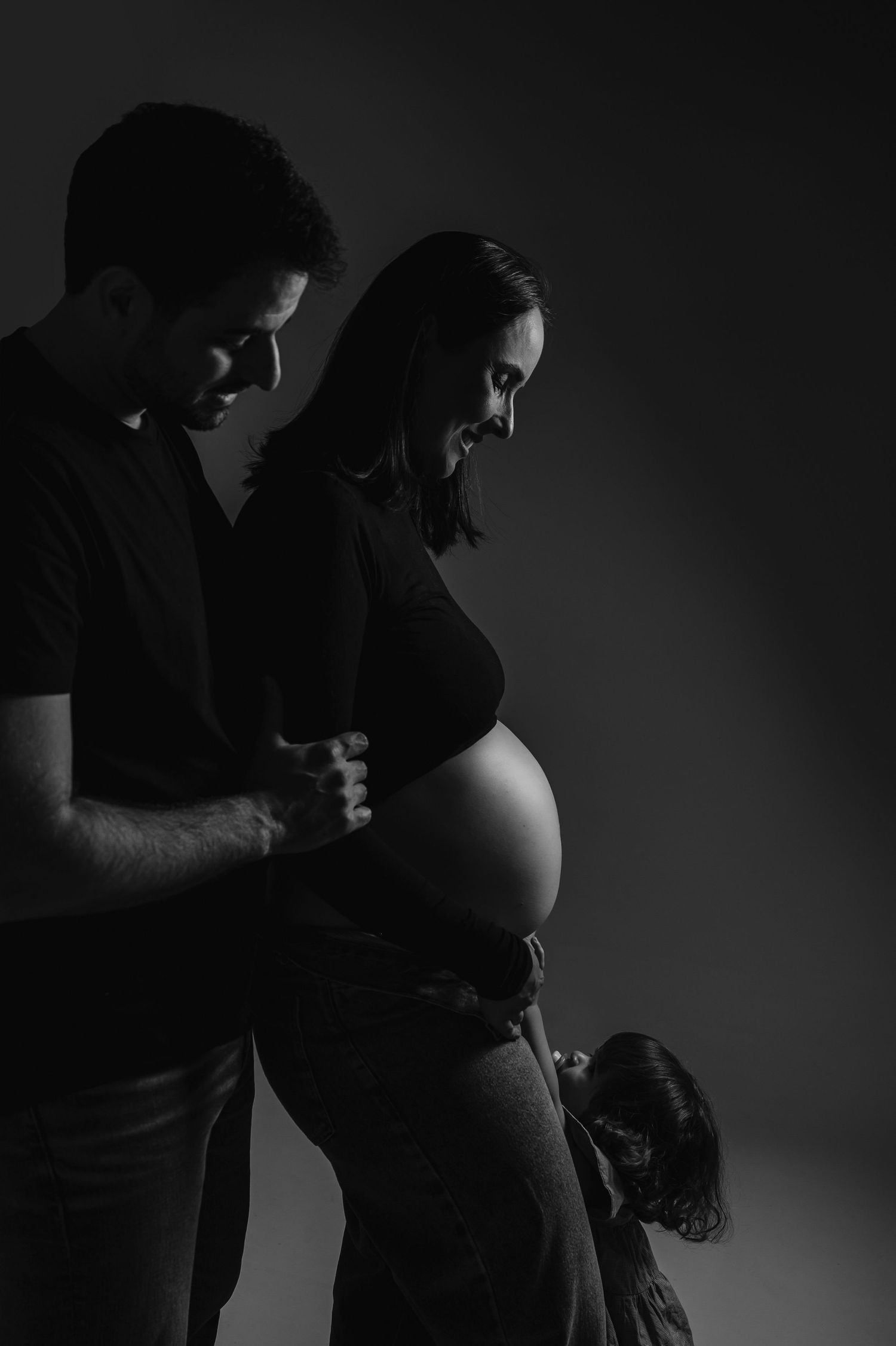 Black and white maternity photo of couple and child tenderly touching pregnant belly in dramatic studio lighting.