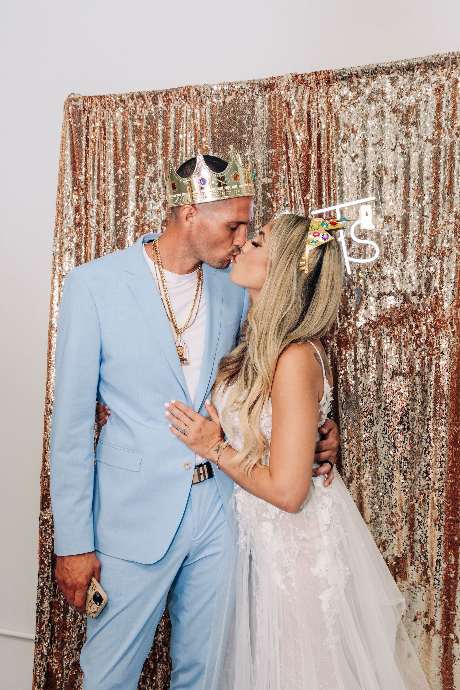 A couple in light blue suit and white wedding gown share a kiss in front of a rose gold sequin backdrop while wearing crowns in Lantana.