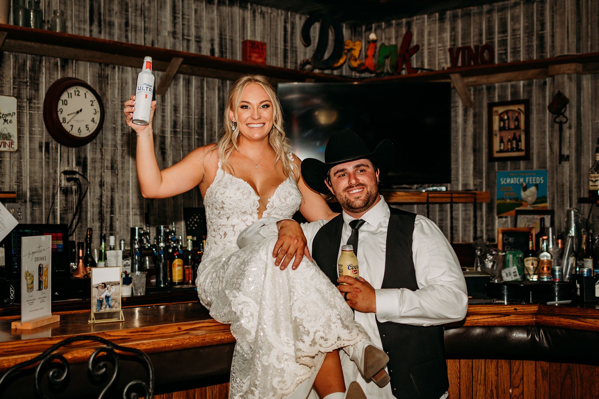 Bride and groom celebrating at a rustic bar, bride holding a bottle aloft in a wedding dress while groom kneels beside her.
