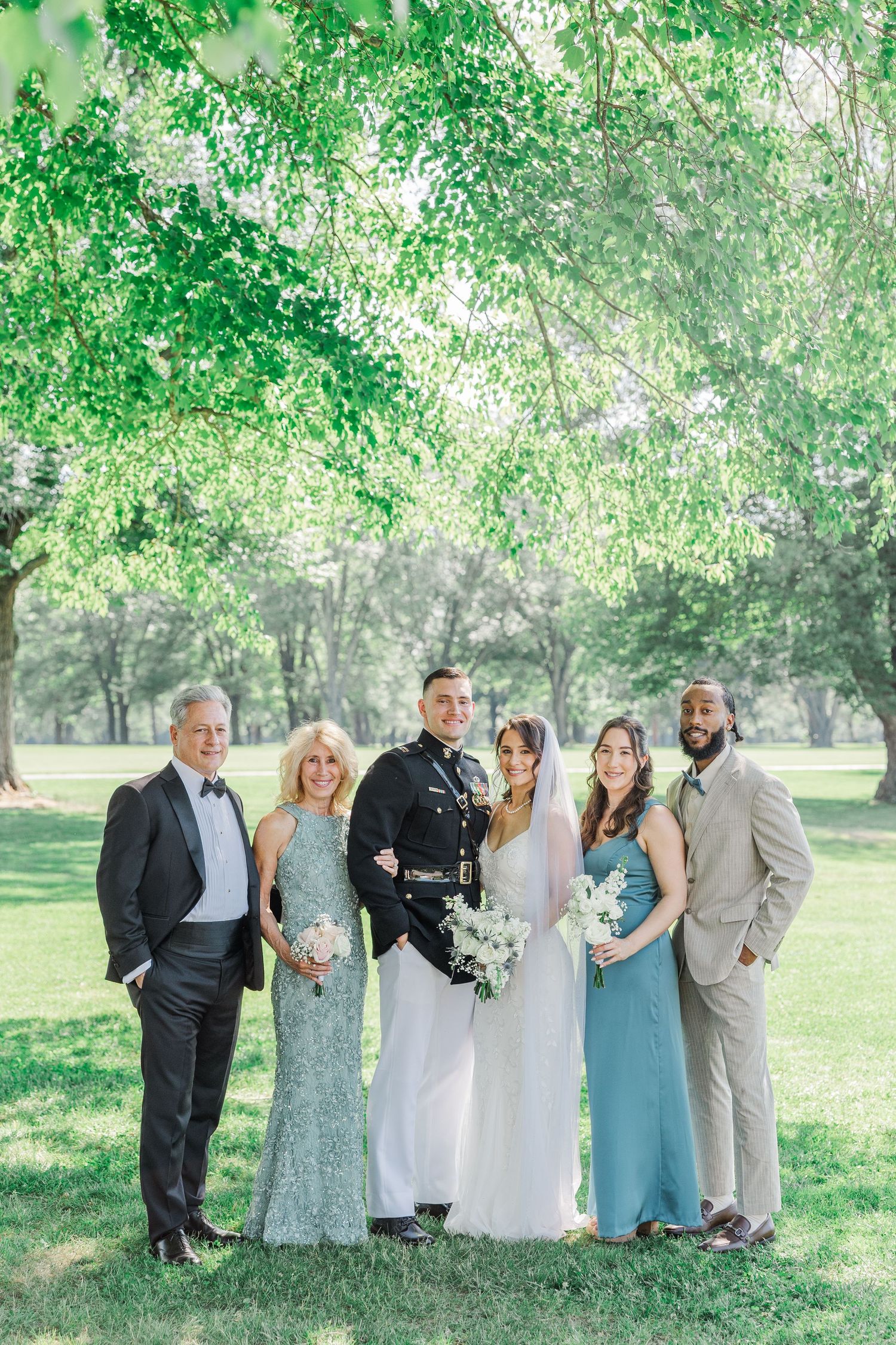Military groom and bride pose with wedding party in elegant attire outdoors under lush green trees on a sunny day.