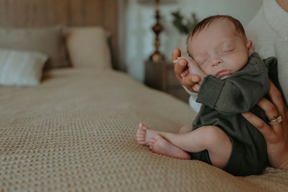 Newborn baby sleeping peacefully in dark green romper on a beige knit blanket beside a bed.