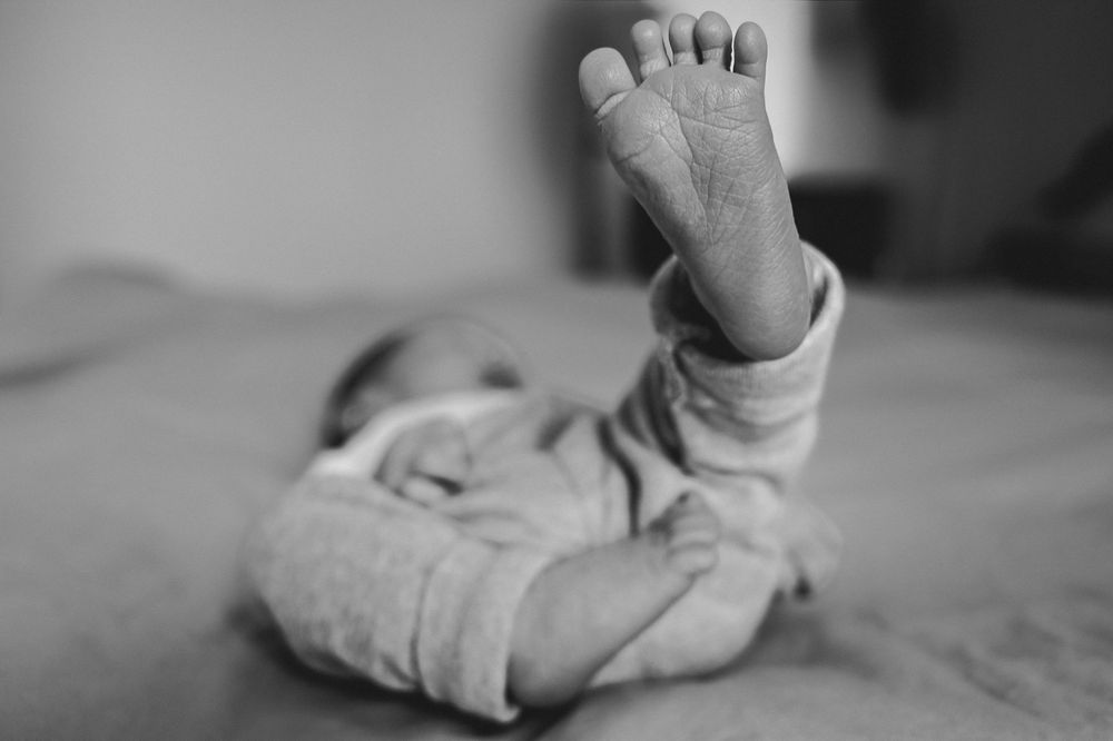 Black and white photo of a newborn baby lying on a bed with tiny foot raised in the air.
