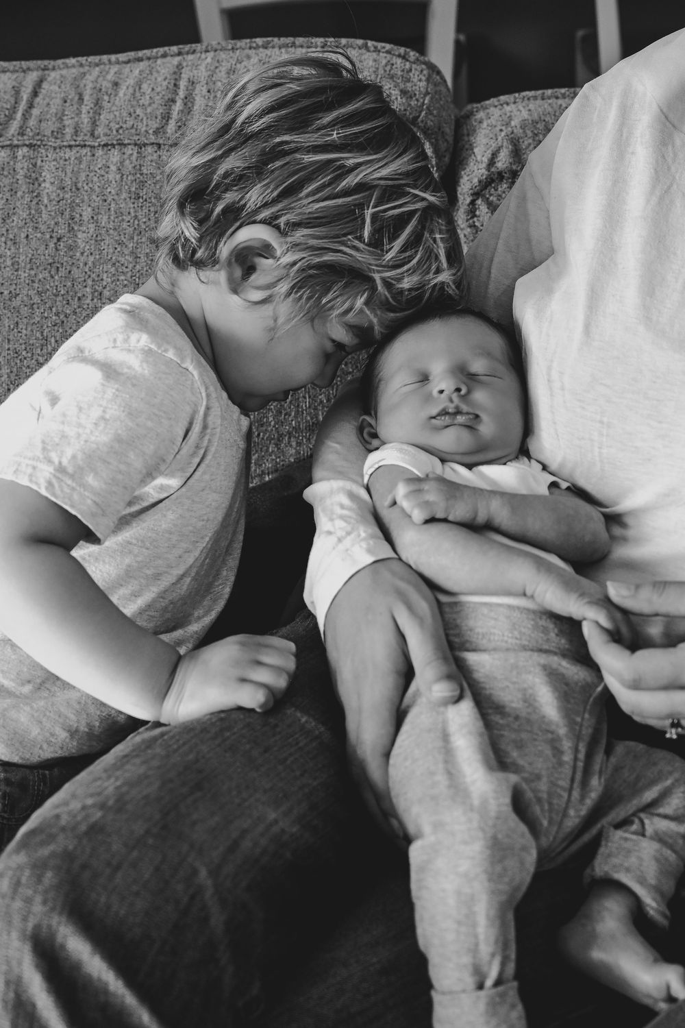 Black and white photo of young child leaning over sleeping newborn baby held by adult on couch.