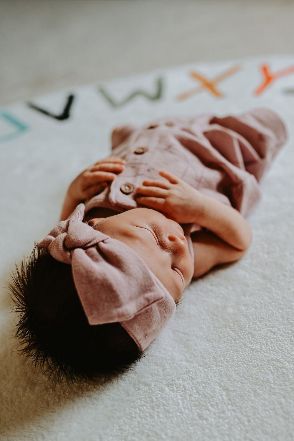 A newborn baby in a pink outfit and headband lies on a white alphabet blanket.