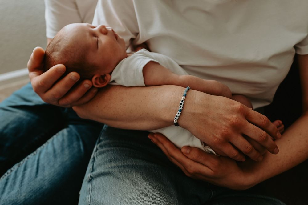 Parent cradling a sleeping newborn baby in their arms, wearing a bracelet, dressed in blue jeans.