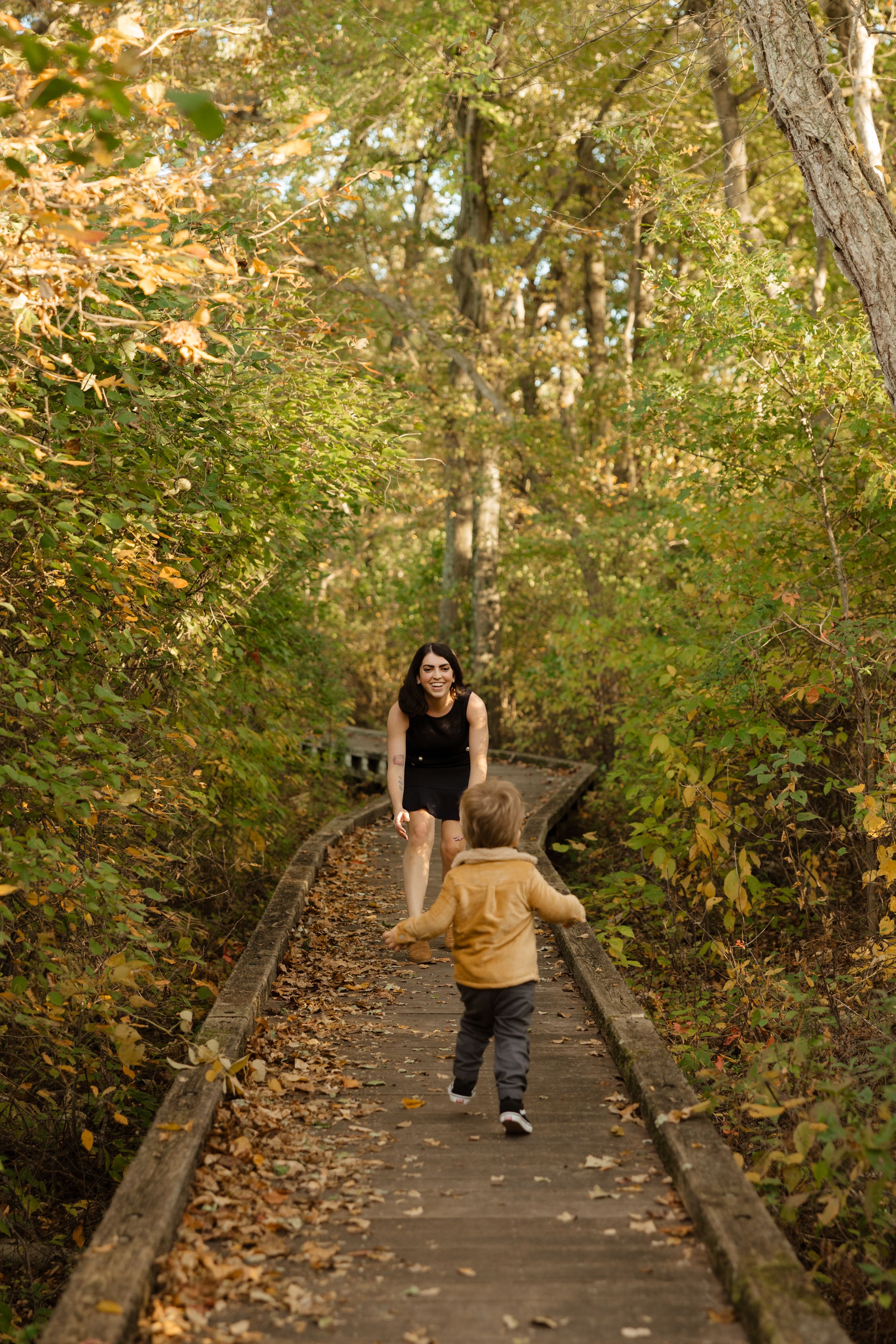 Child running toward smiling woman on wooden boardwalk trail surrounded by colorful autumn foliage.