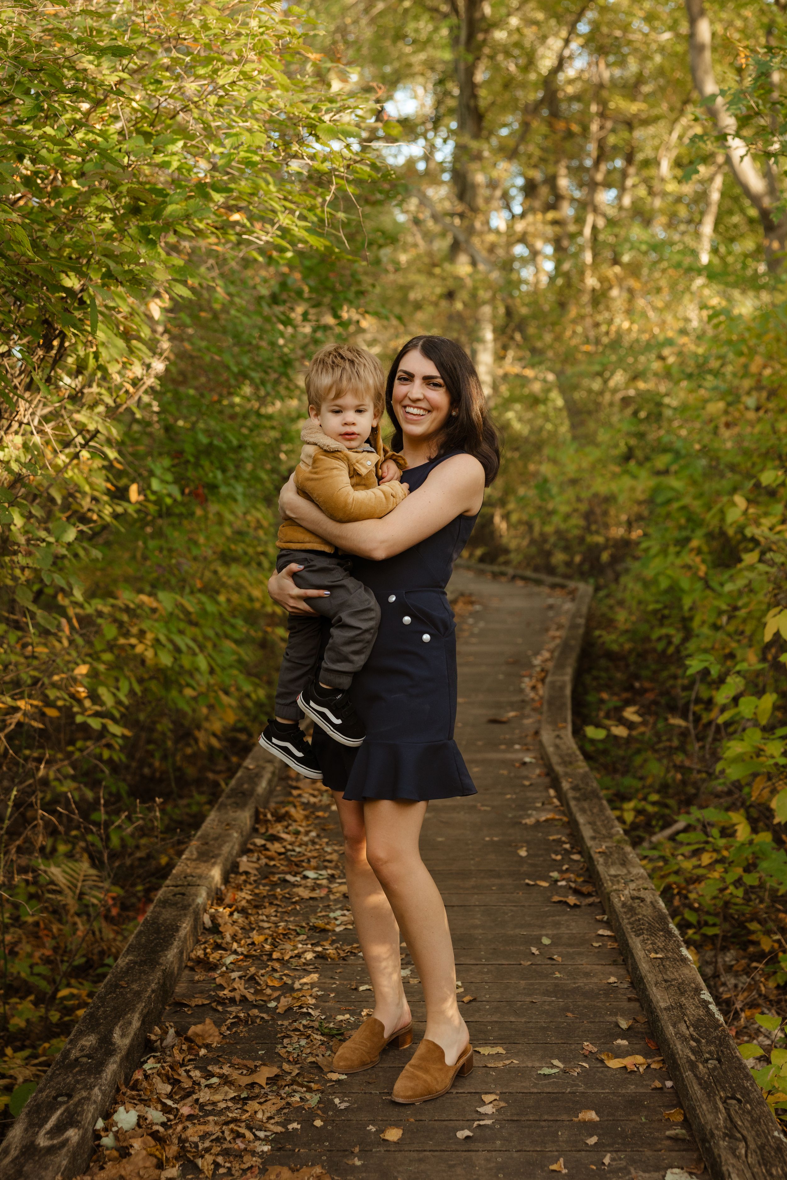 A smiling woman in a navy dress holds a toddler on a wooden boardwalk path surrounded by autumn foliage.