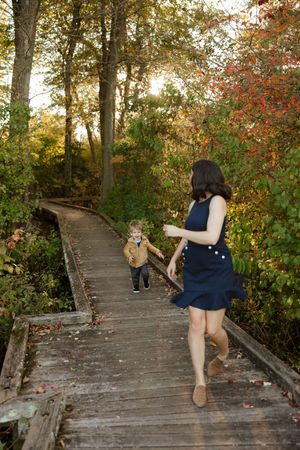 A mother in a navy dress chases her toddler on a wooden boardwalk path surrounded by colorful autumn foliage.