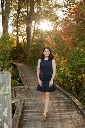 Young woman in navy dress walks wooden boardwalk path surrounded by autumn foliage with golden sunlight.