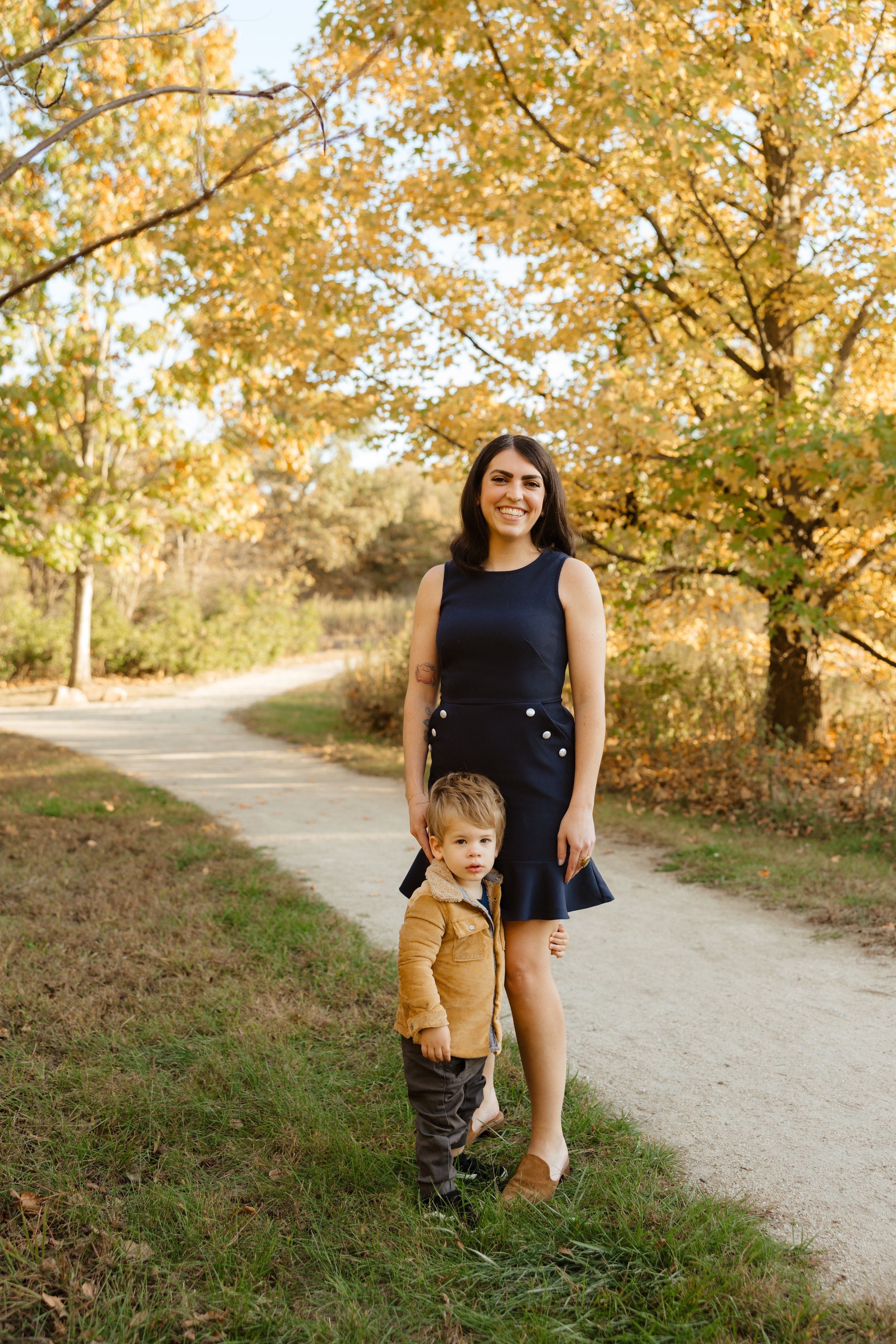 A smiling woman in a black dress stands with a toddler in a yellow jacket on a scenic autumn park path.