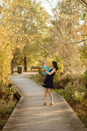 Mother in black dress holds toddler on sunny autumn boardwalk path surrounded by golden fall foliage.