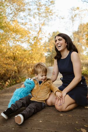 A laughing woman and toddler boy sit on a wooden path in autumn, the child holding a blue Cookie Monster plush toy.