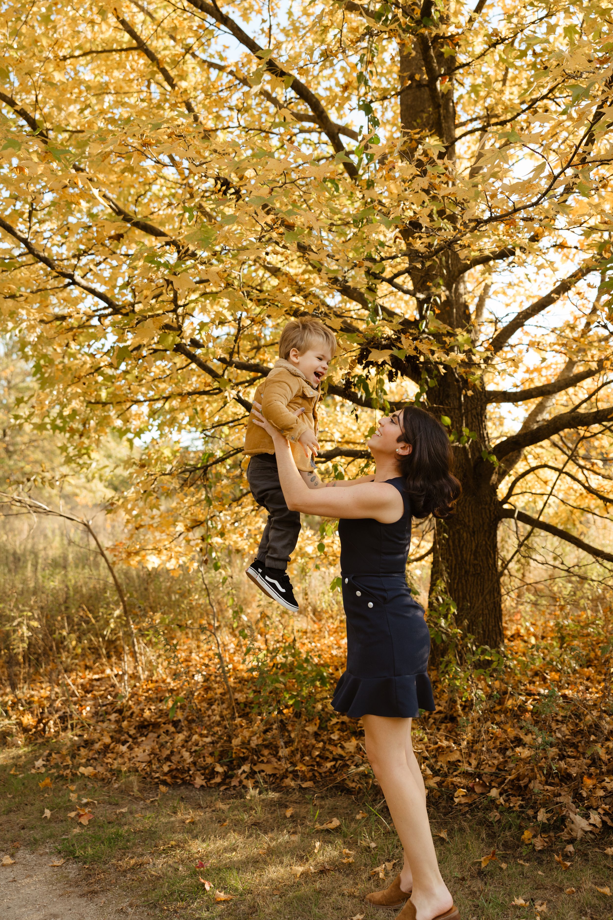 A joyful mother lifts her laughing toddler under golden autumn trees in a park.