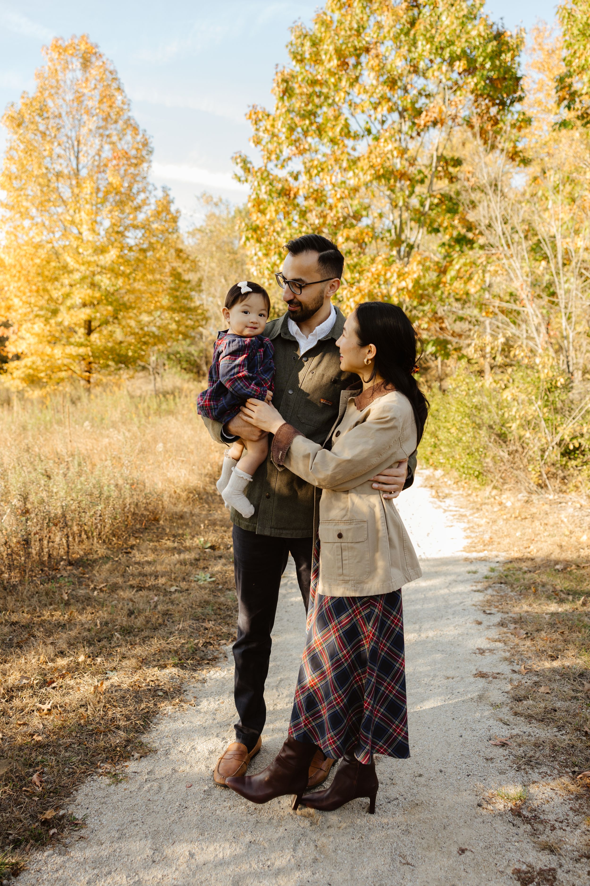 Family of three poses on a autumn trail, father holding baby girl, mother beside them, surrounded by golden fall foliage.