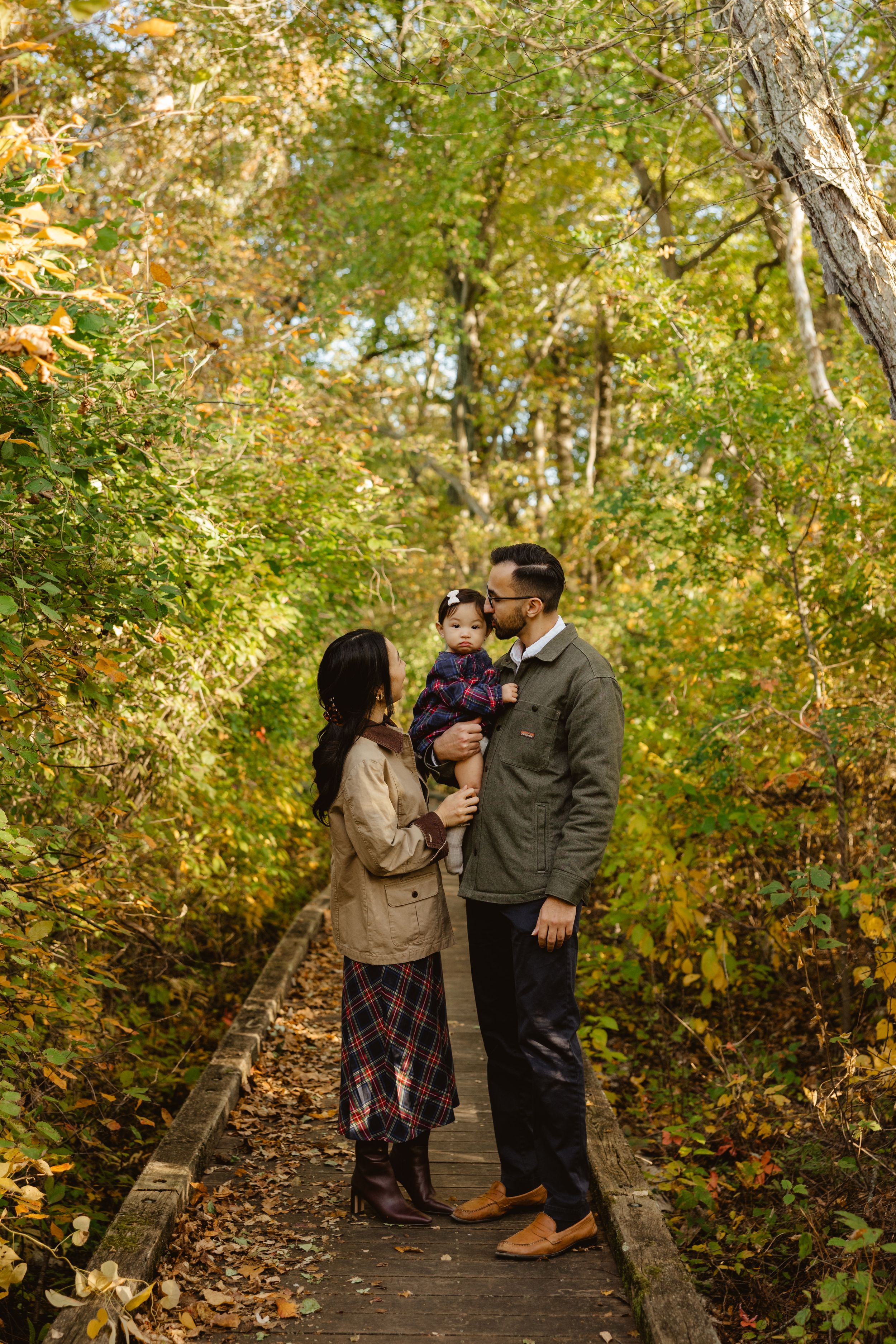 Family of three poses on a wooden boardwalk trail surrounded by colorful autumn foliage on a sunny fall day.