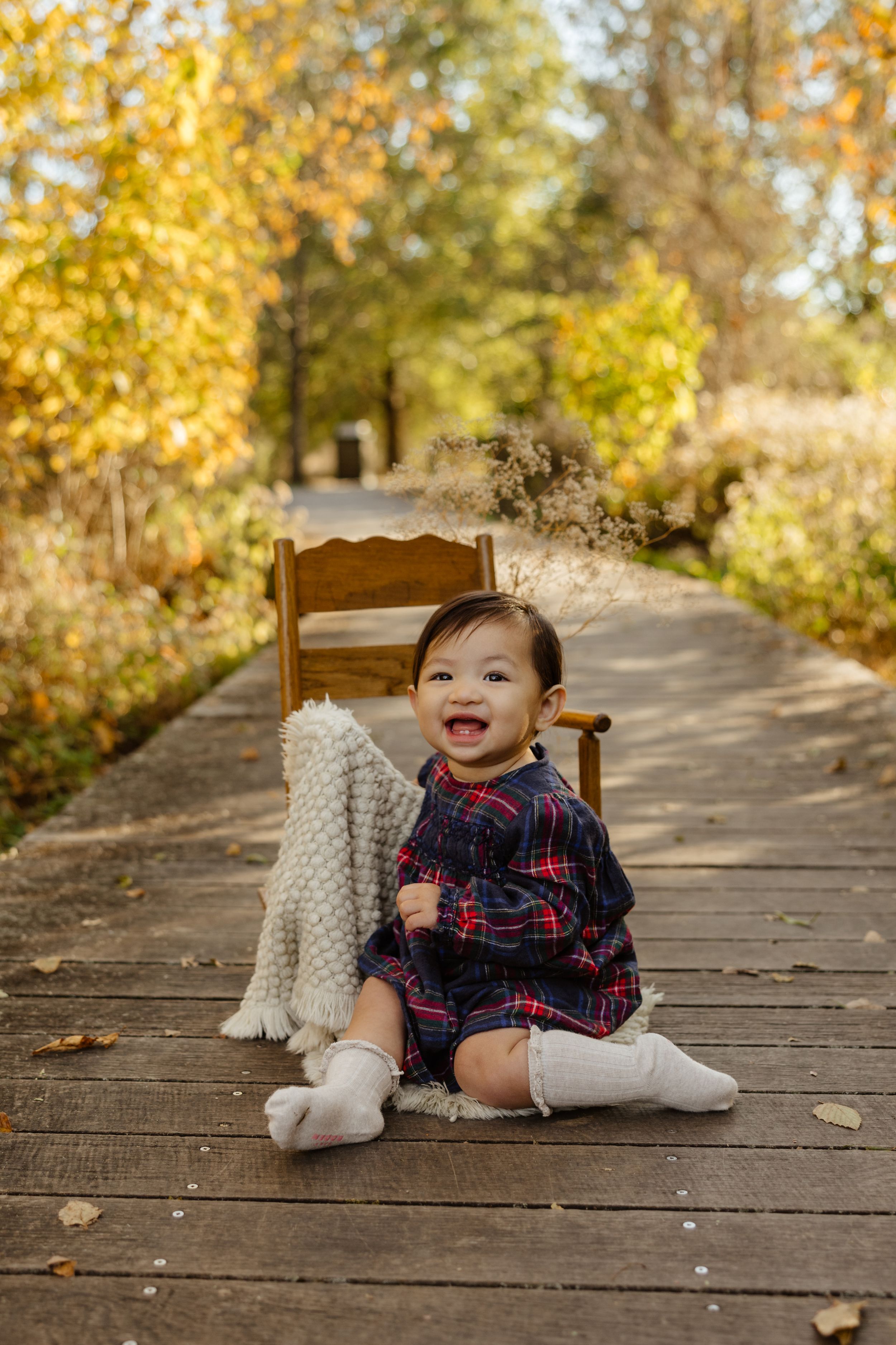 Smiling baby in plaid dress and white socks sitting on wooden boardwalk surrounded by golden autumn foliage.
