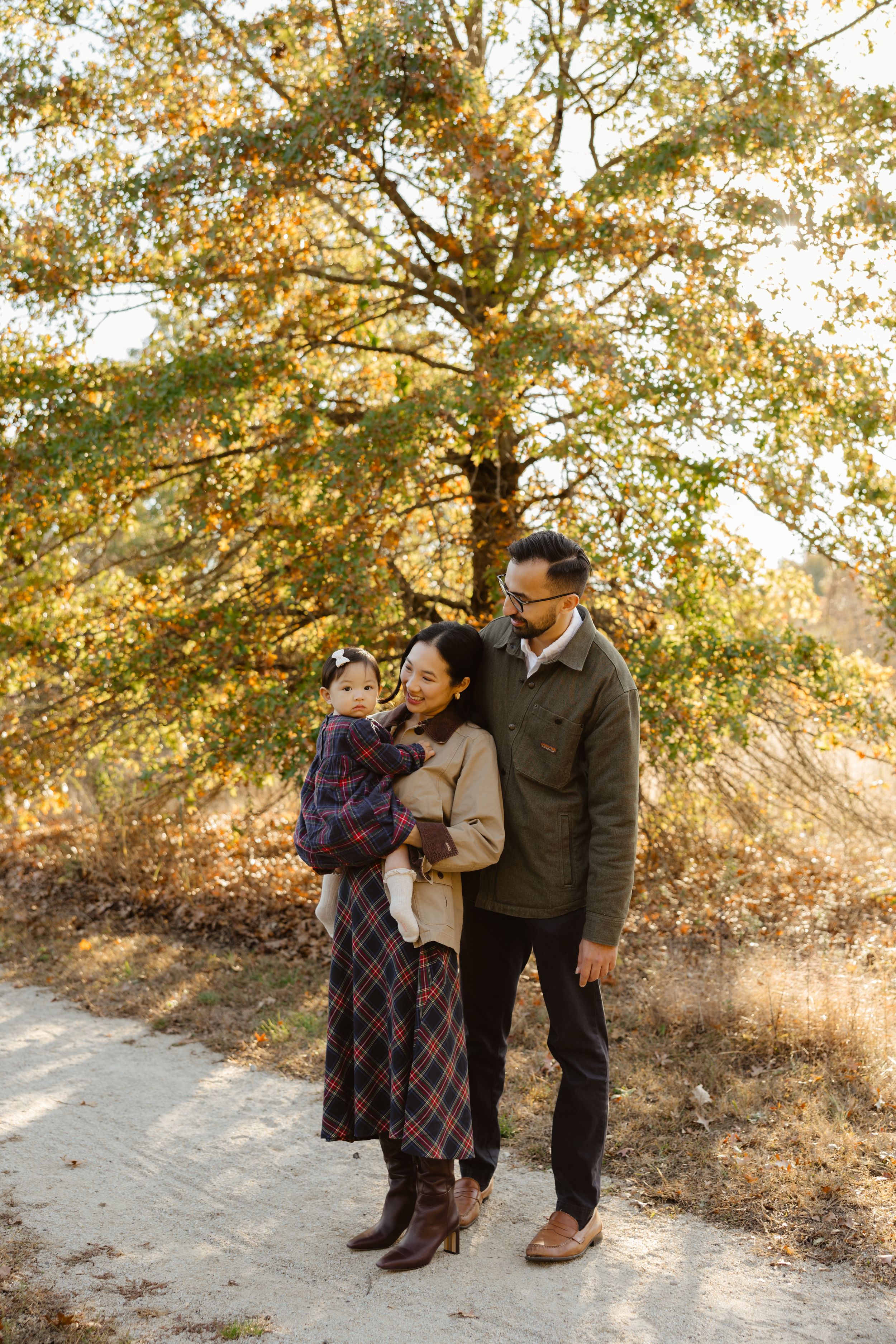 A young Asian family poses outdoors in fall, parents holding toddler near golden autumn tree on a sunlit path.