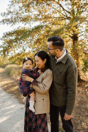 A happy Asian family with a baby girl poses outdoors on a path surrounded by autumn foliage.