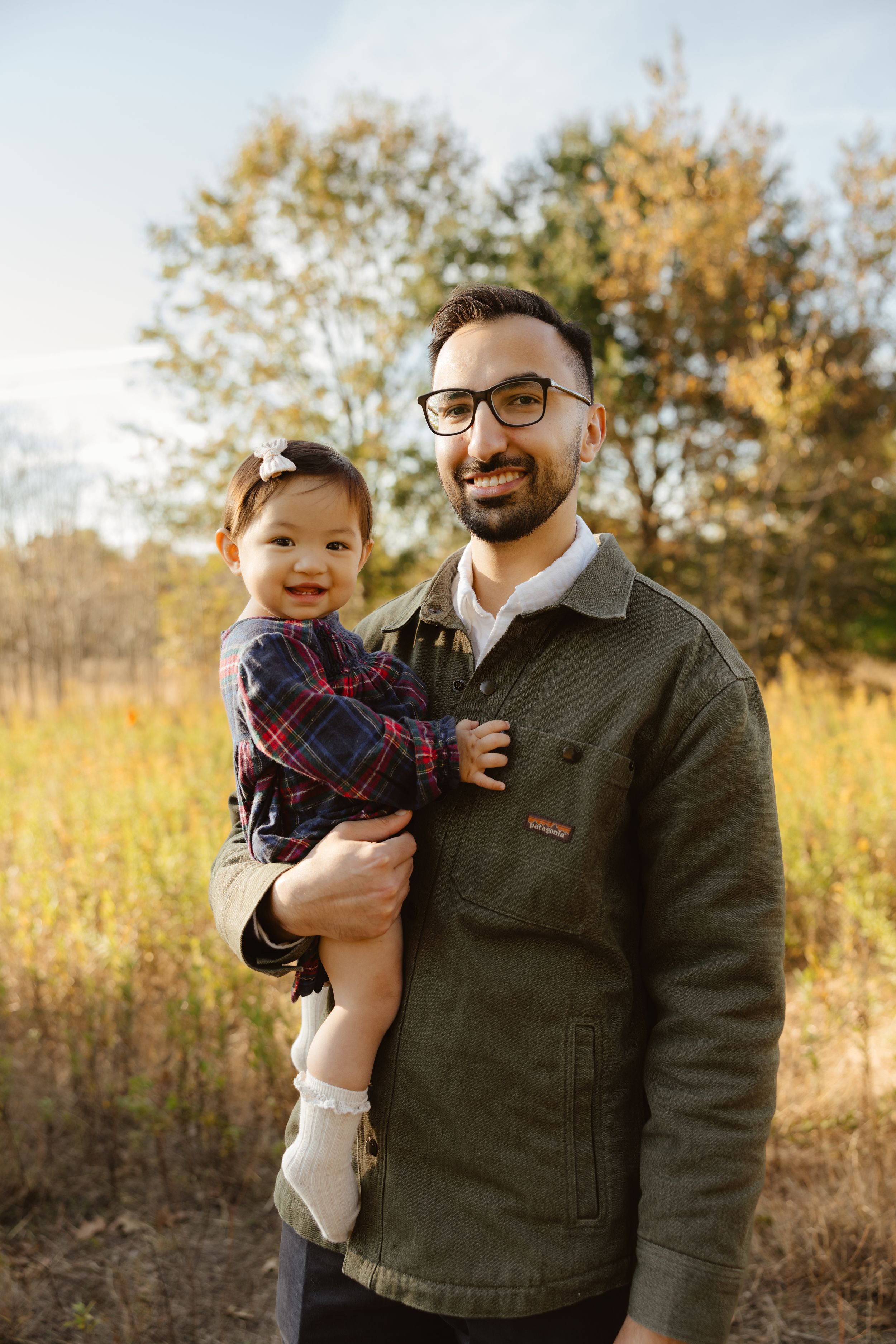 A smiling man with glasses holds a baby girl in a plaid dress outdoors in a sunny autumn field.