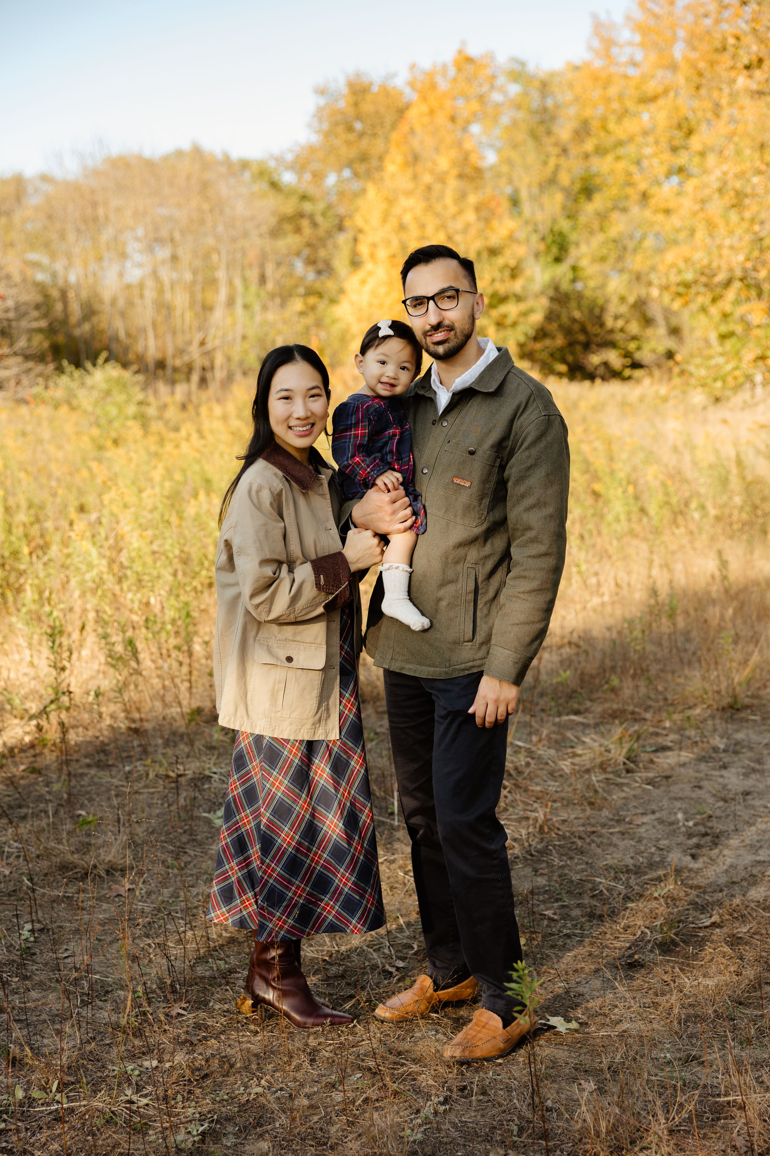 Family of three poses in autumn field, parents holding toddler, surrounded by golden fall foliage.