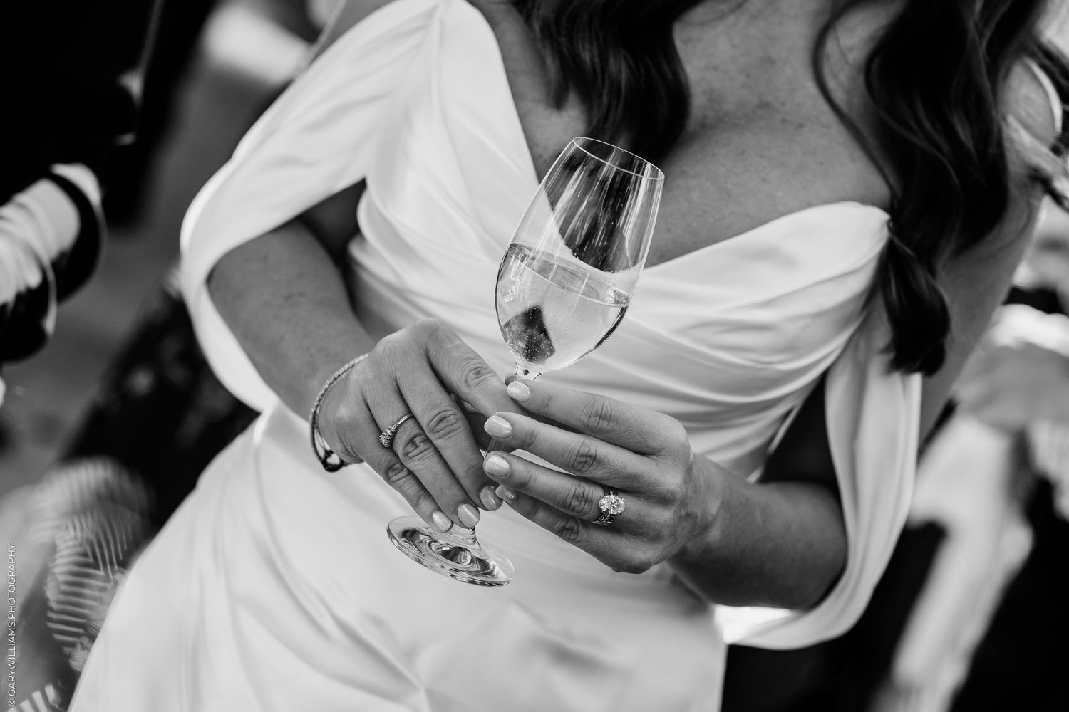 Black and white photo of a bride in white gown holding a champagne glass, showcasing elegant jewelry and manicured nails.