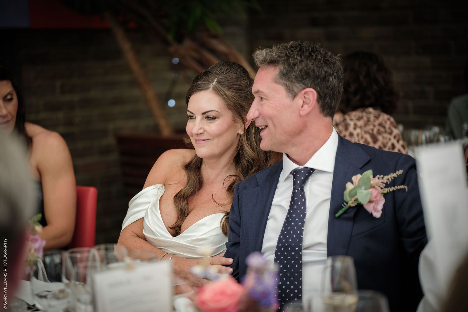 Bride and groom share a tender moment at wedding reception dinner table, bride in white gown, groom in navy suit.