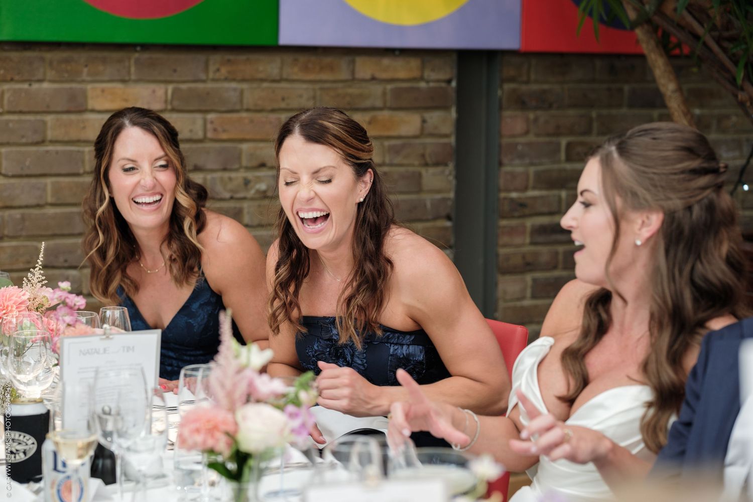 Three bridesmaids laughing joyfully at a wedding reception table with floral centerpieces and colorful banners.