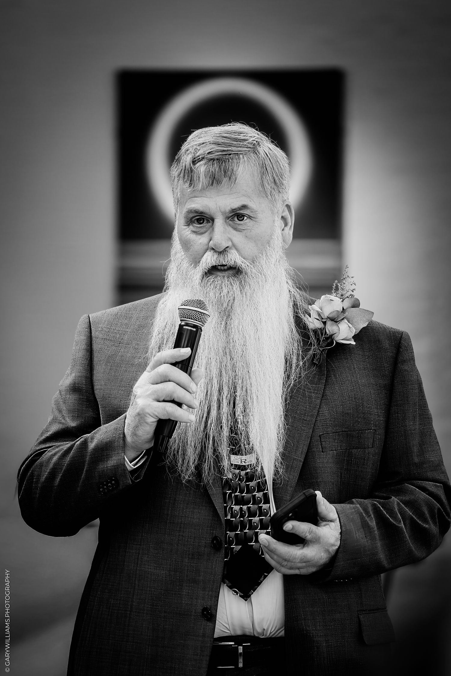 Elderly man with long white beard speaking into microphone at formal event wearing suit with boutonniere.