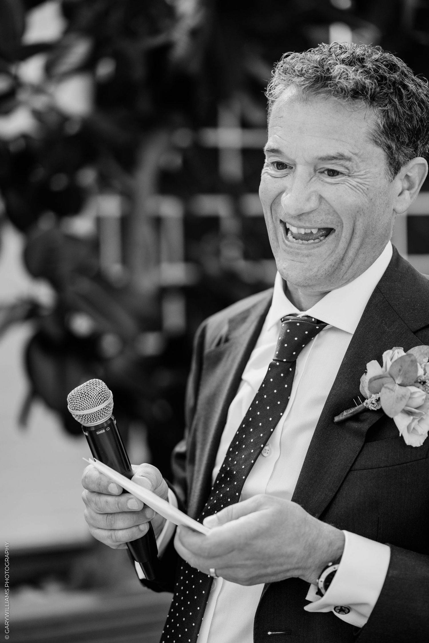 Man in suit with boutonniere smiling while giving a speech holding microphone and notes at a wedding reception.