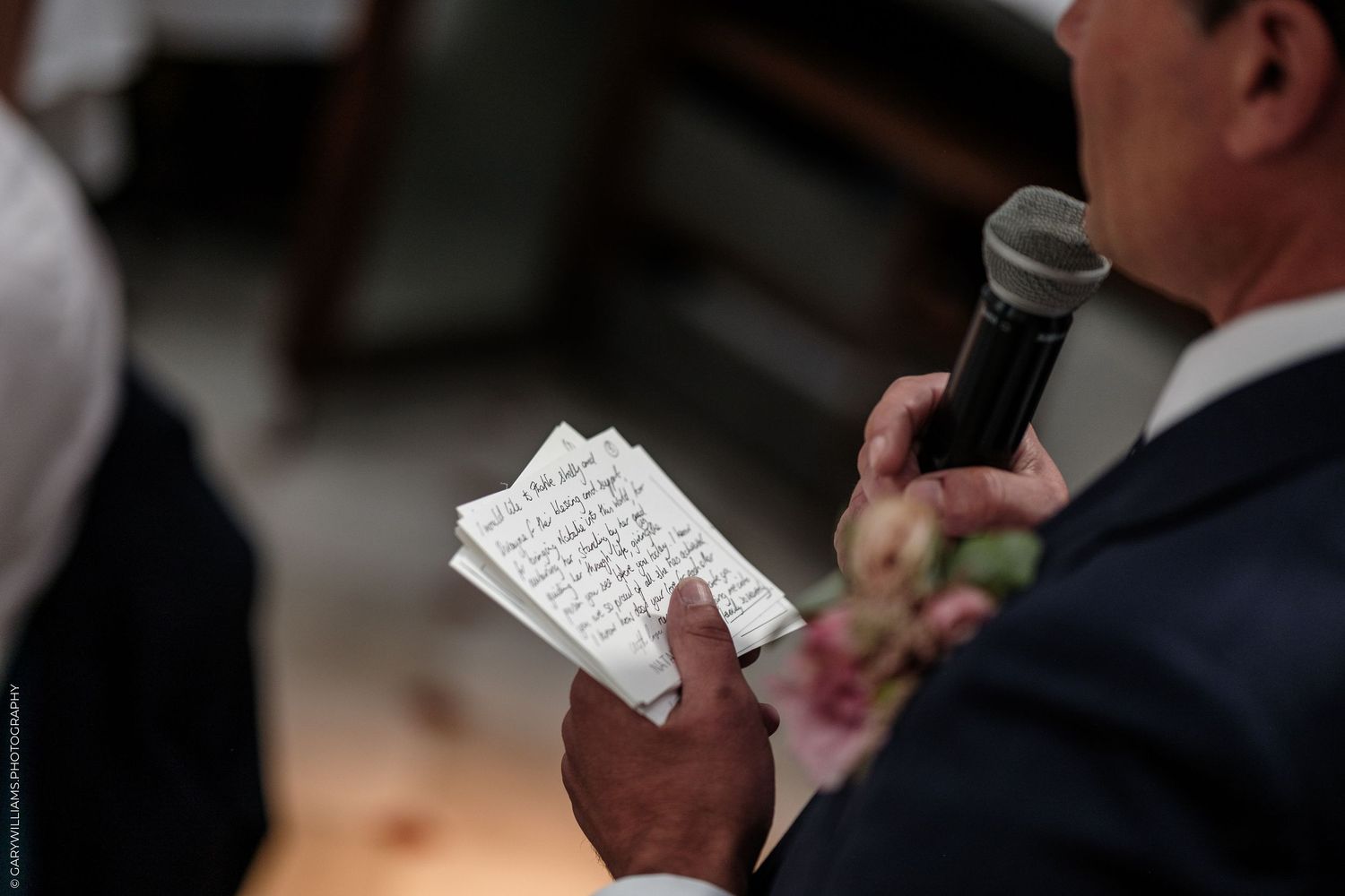 Man in suit holding handwritten notes and microphone during wedding speech ceremony.