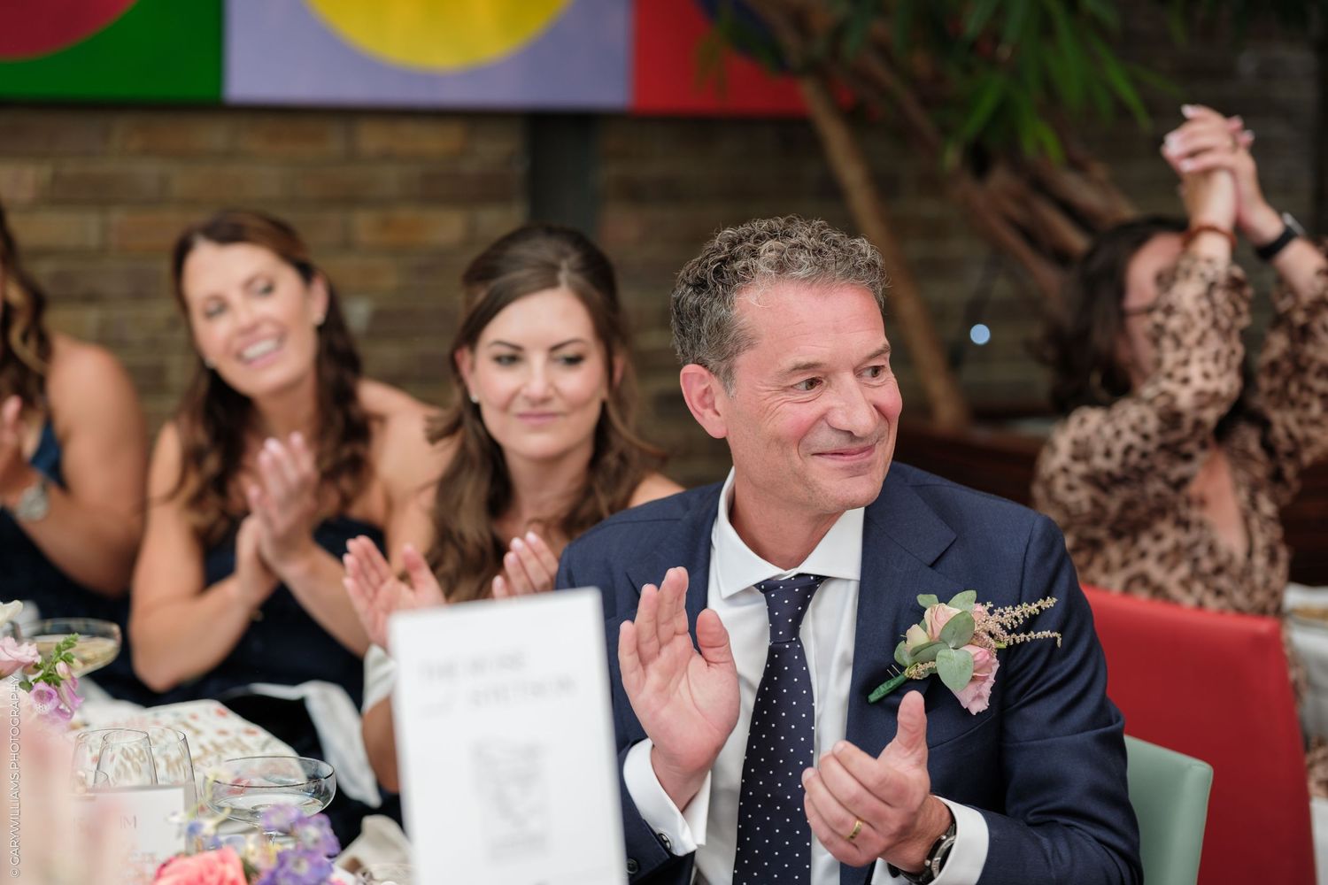 Man in navy suit with boutonniere clapping at wedding reception, bridesmaids smiling in background.