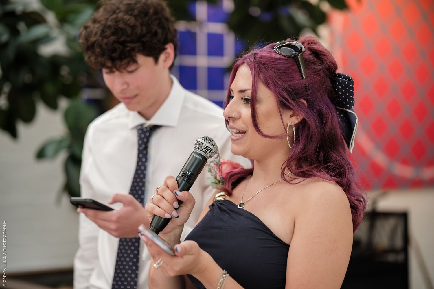 Woman with purple hair singing into microphone while holding wine glass at indoor event.