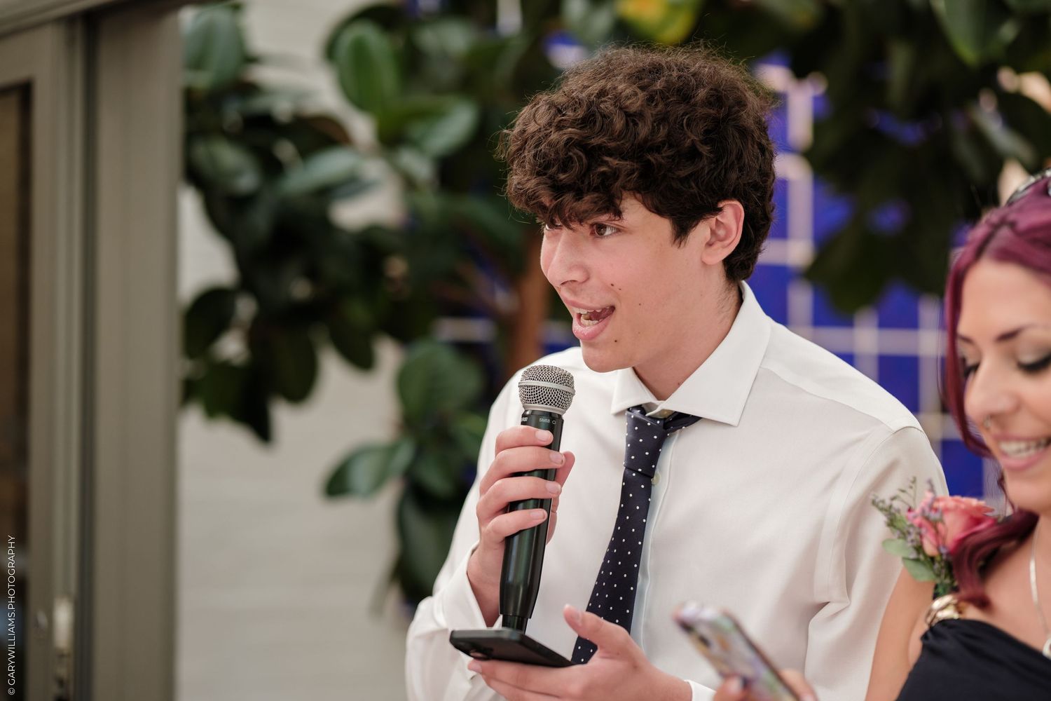 Young man in white shirt and tie speaking into microphone at formal event with woman beside him.