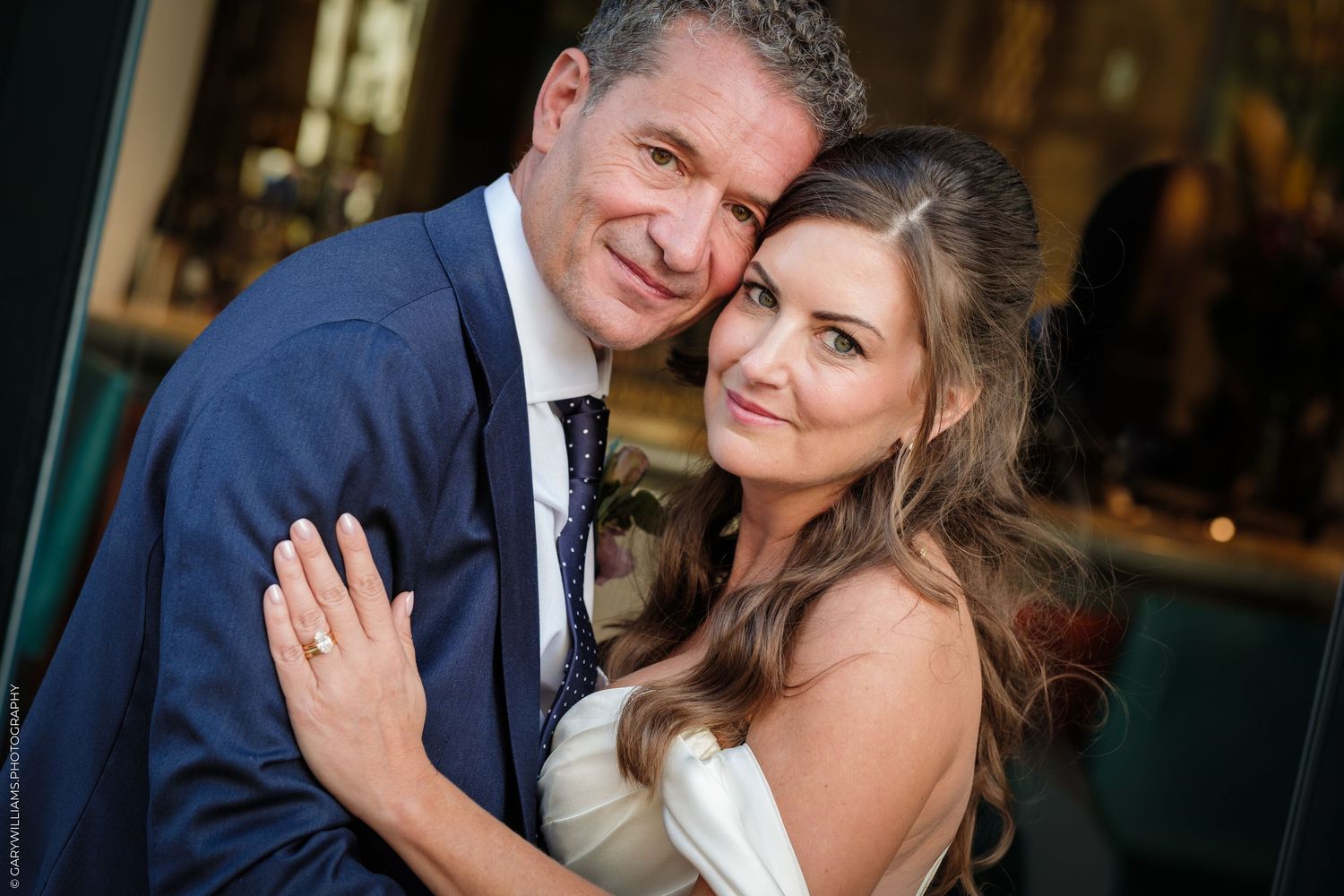 Smiling bride and groom pose closely together at their wedding reception, she wearing white and showing her ring.