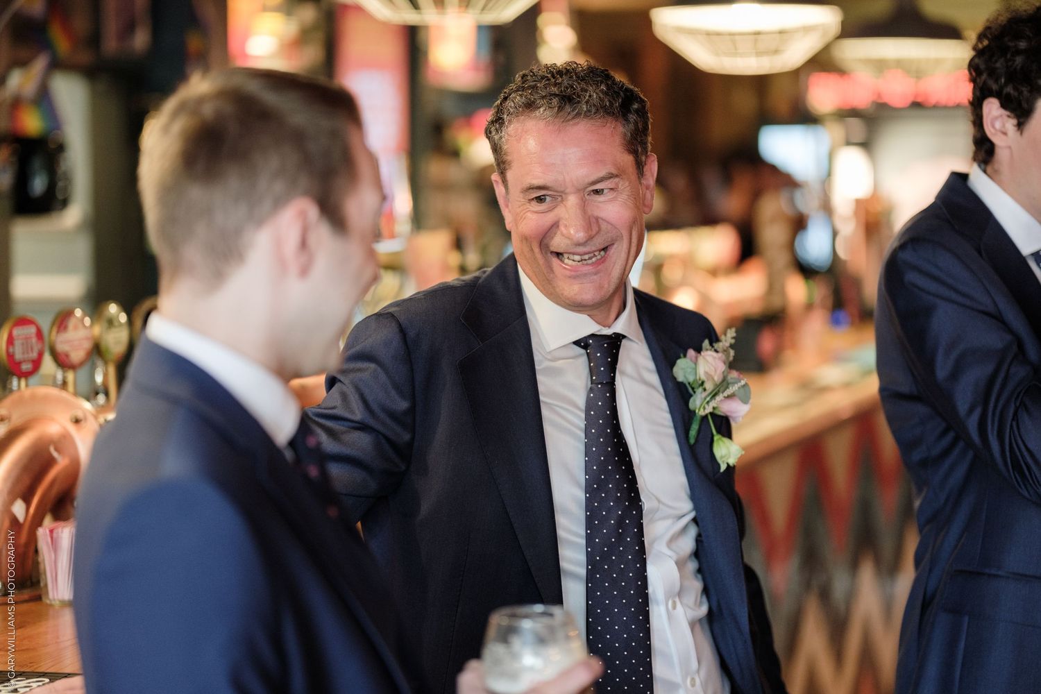 Men in suits socializing at a wedding reception bar, smiling and holding drinks in an elegant venue.
