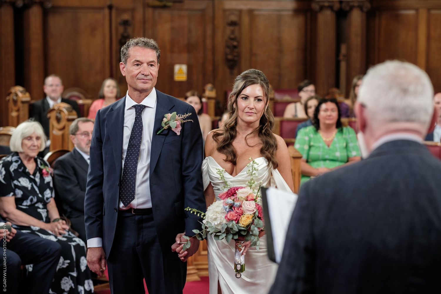 Bride and groom hold hands at wedding ceremony altar in elegant wood-paneled venue with guests seated behind.
