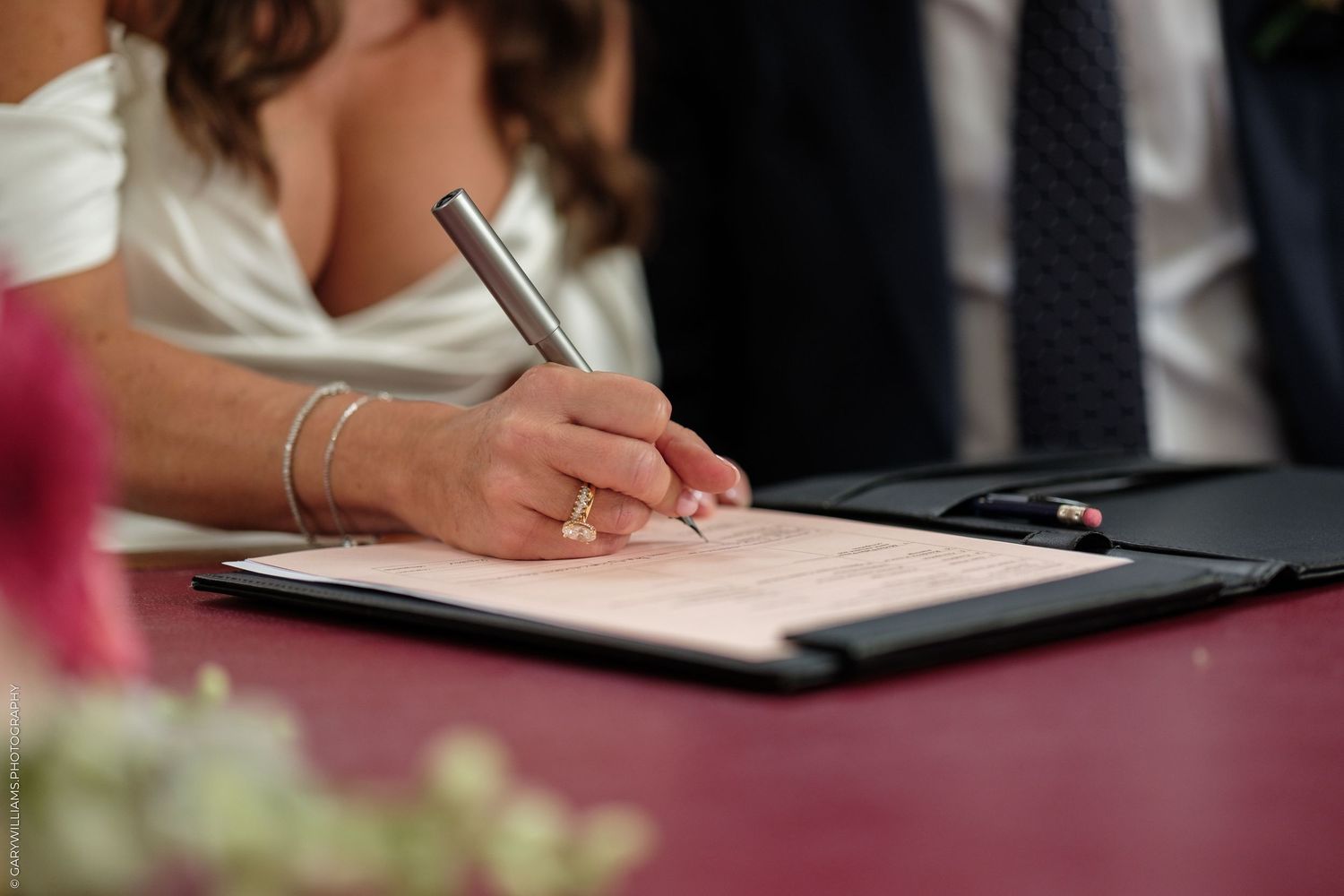 Bride signing wedding certificate with elegant pen, wearing bridal gown and gold bracelet at ceremony table.