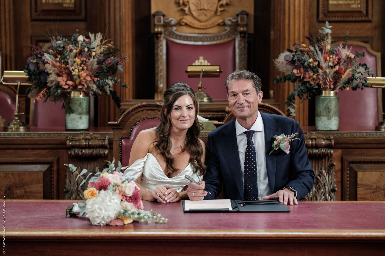 Couple smiling at a wedding ceremony table with floral arrangements in an ornate wood-paneled room.