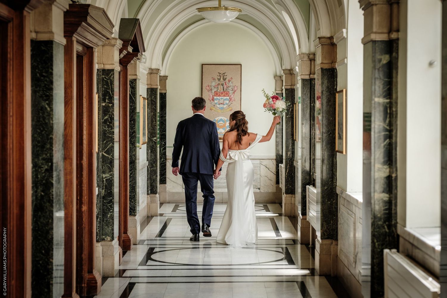 Bride and groom walking hand in hand down an elegant marble hallway, bride holding a bouquet of flowers.