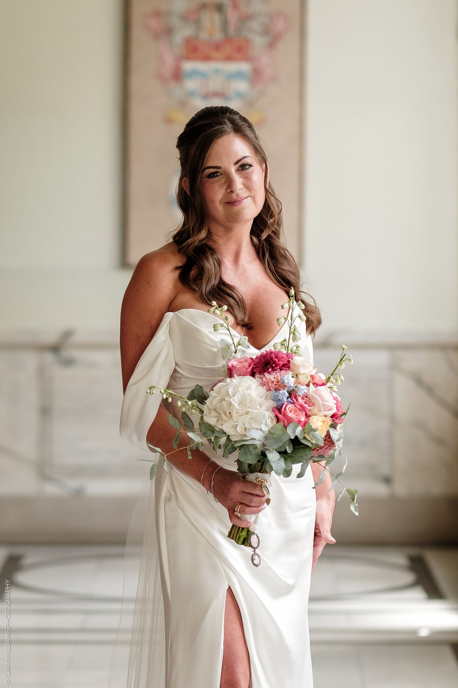 Bride in white off-shoulder gown holding colorful bouquet of pink, white, and blue flowers in elegant marble hall.