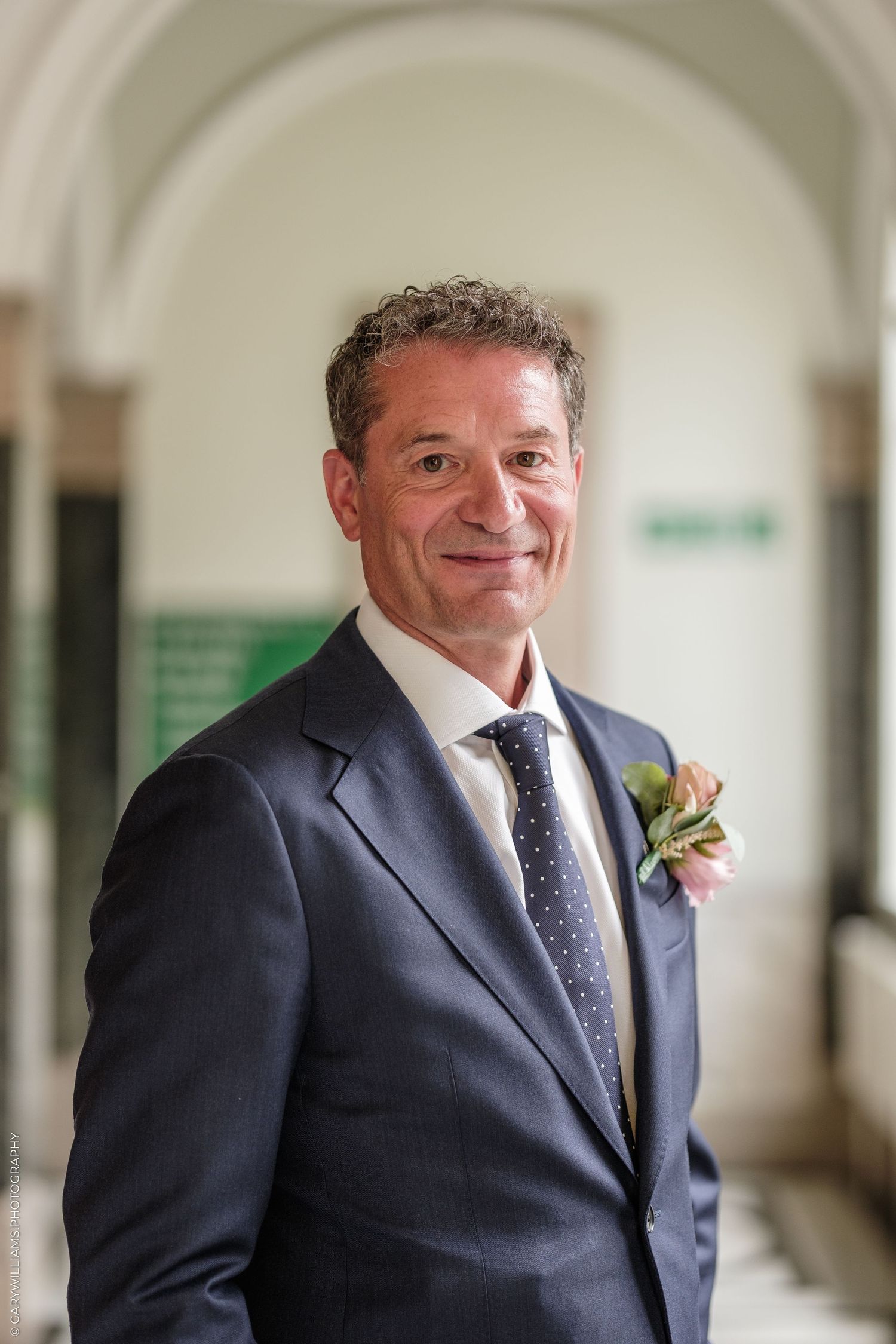 Smiling middle-aged man in navy suit with boutonniere poses in elegant arched hallway.