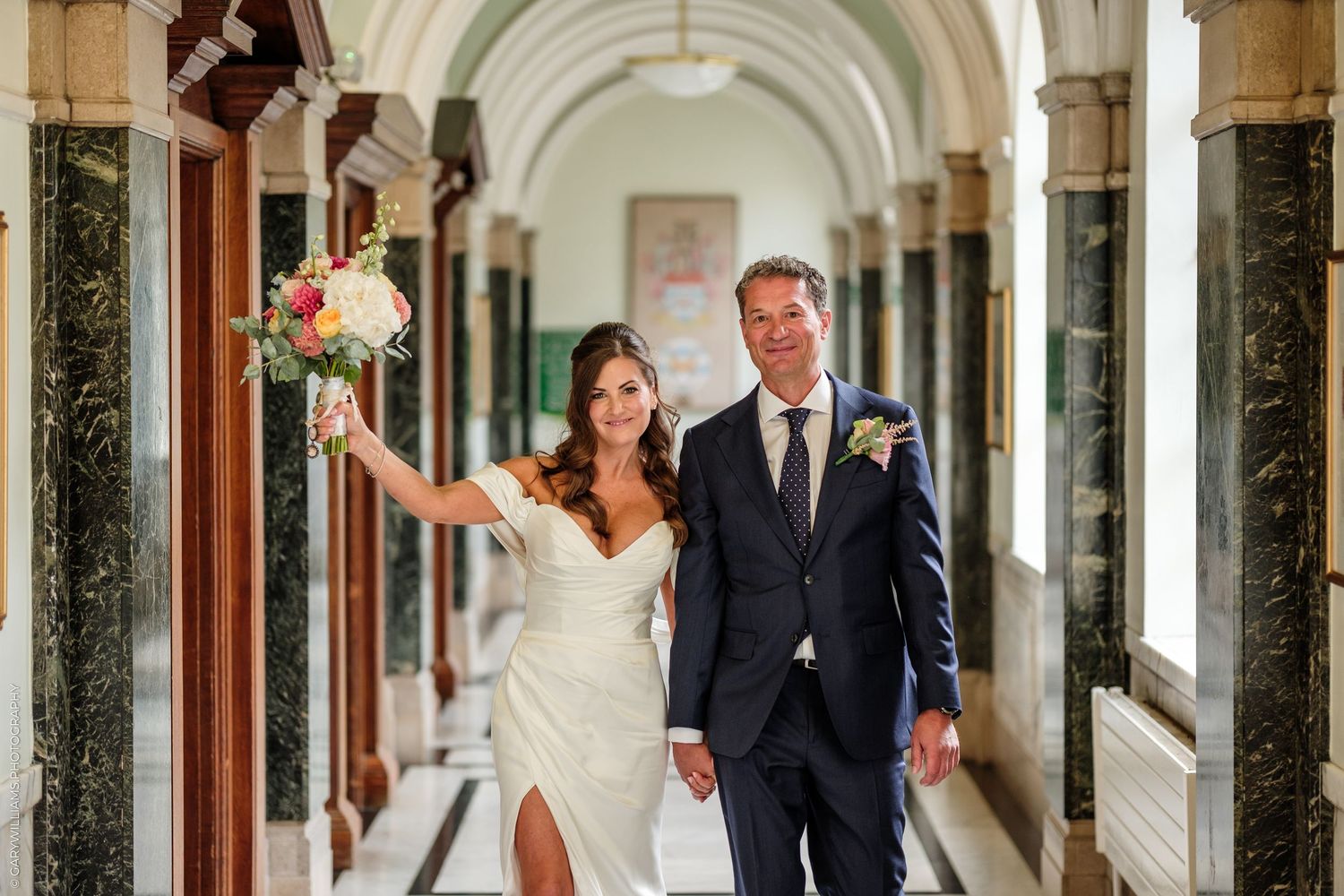Bride and groom walking through elegant marble hallway, bride holding colorful bouquet aloft, both smiling in wedding attire.