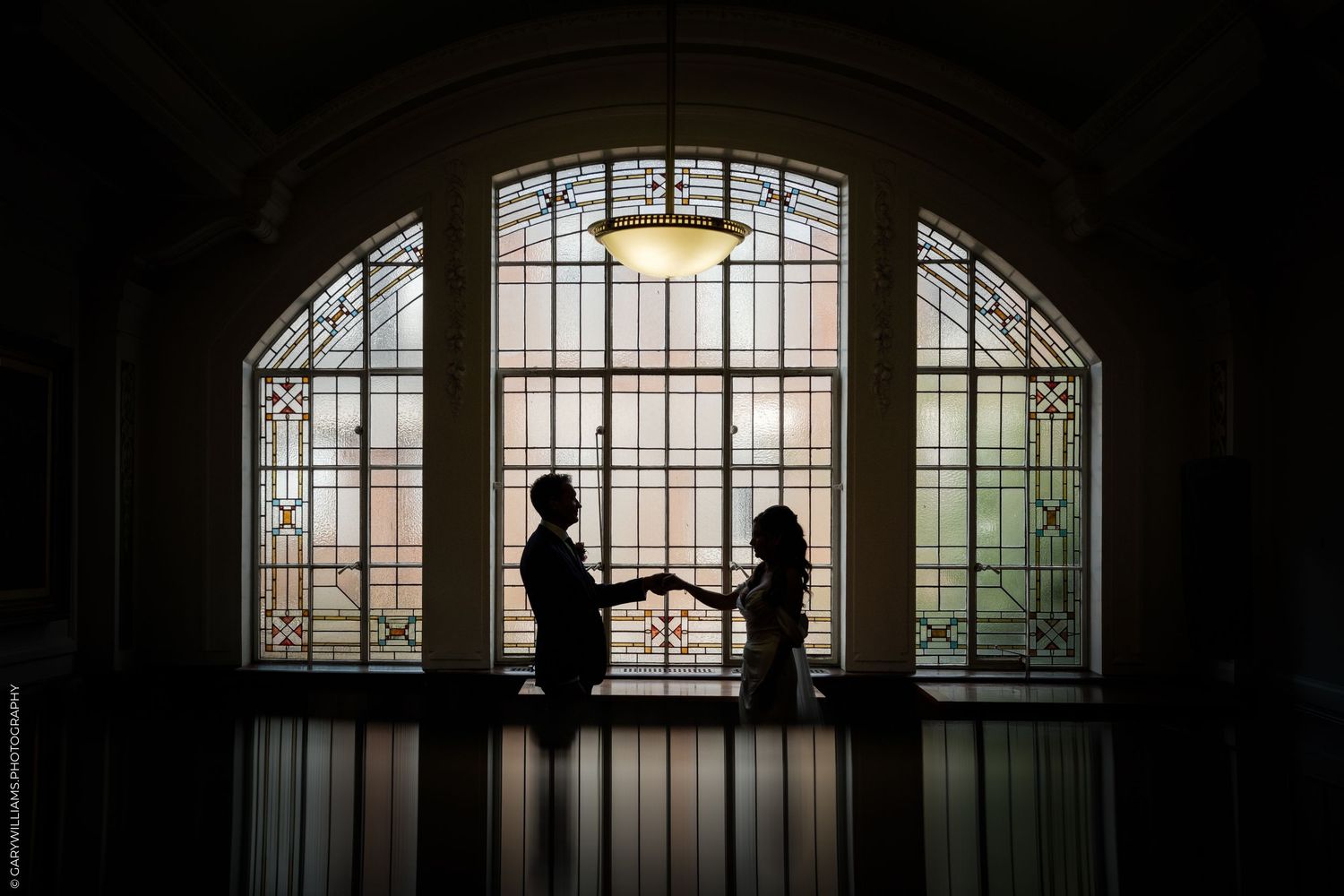 Silhouette of a couple holding hands in front of elegant stained glass windows in a dimly lit venue.