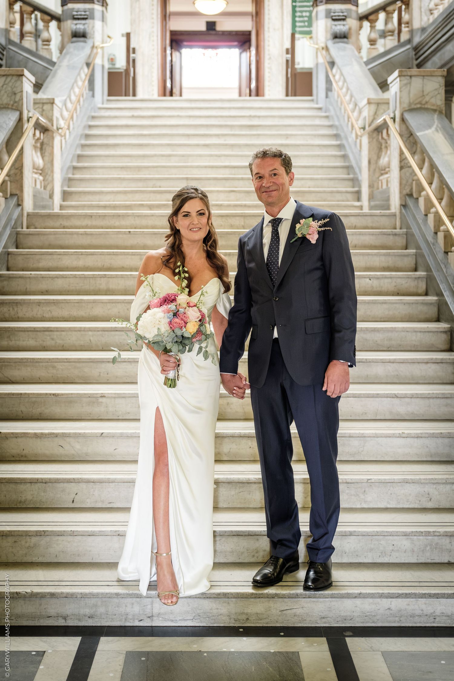 Bride and groom holding hands on marble staircase, bride in white gown with bouquet, groom in navy suit with boutonniere.