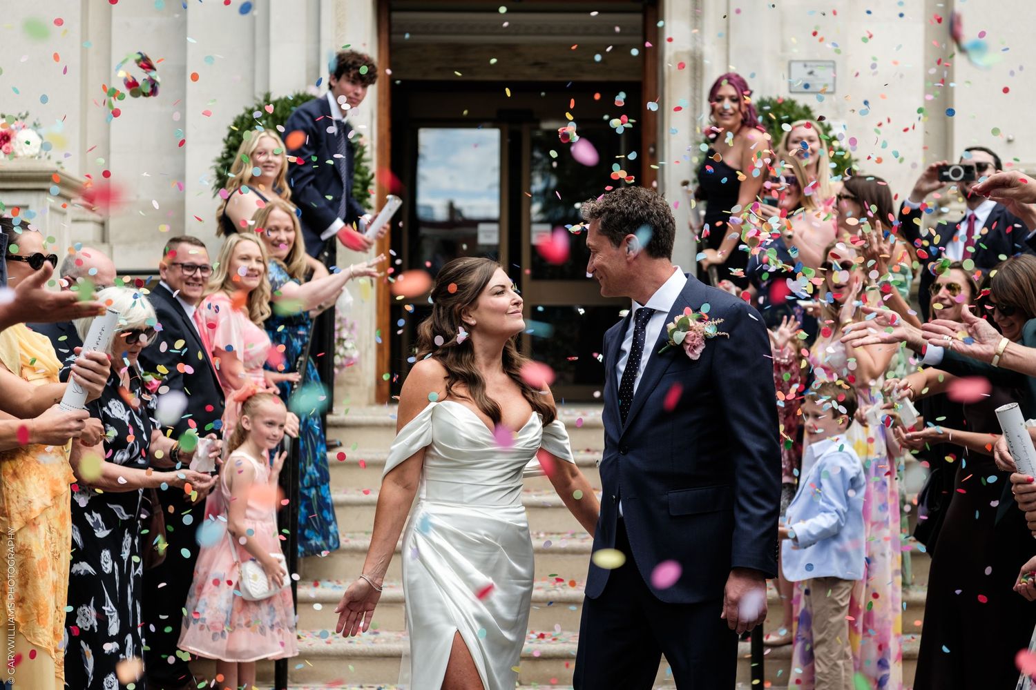 Bride and groom walk through colorful confetti shower outside church surrounded by smiling wedding guests.