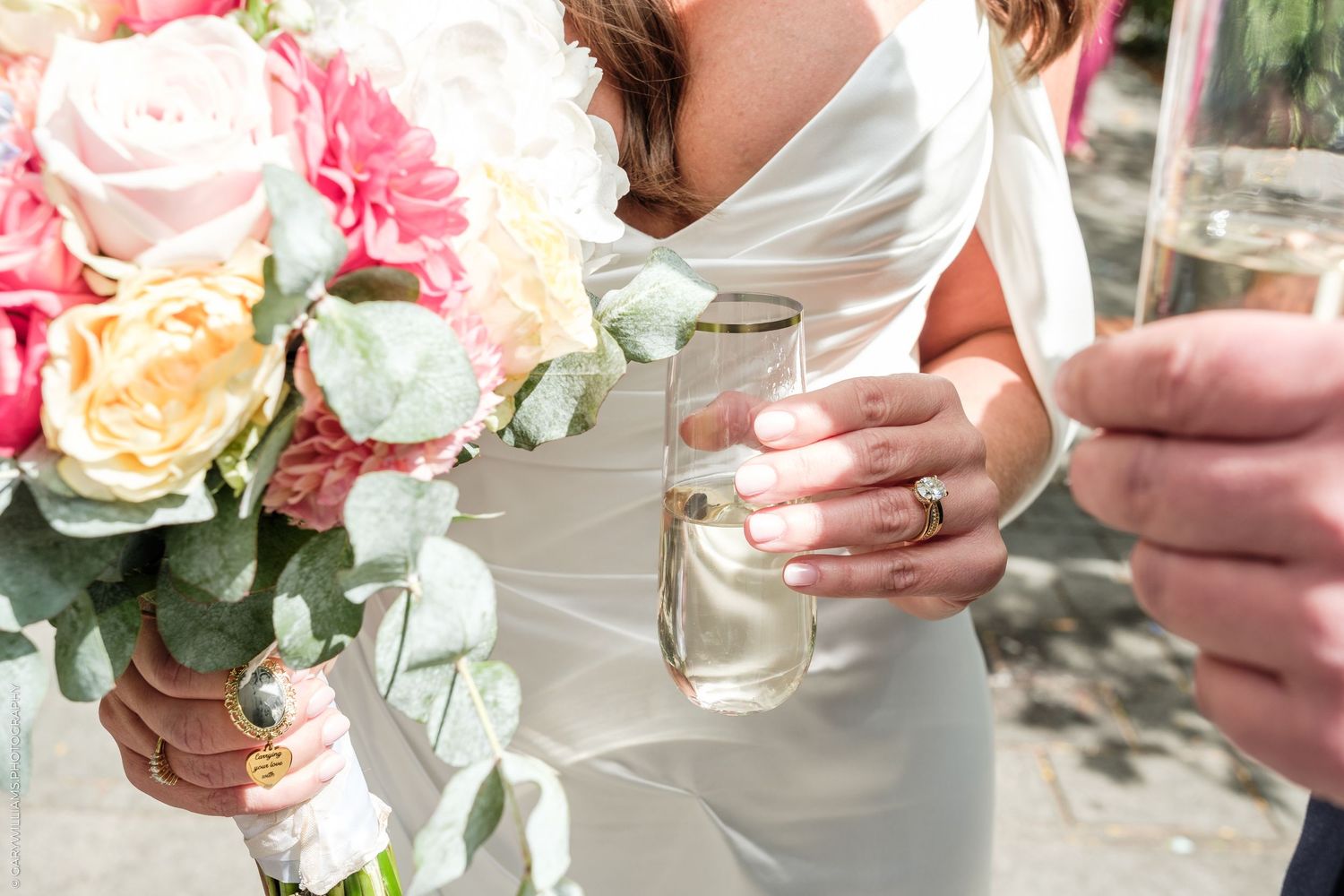 Bride holding colorful pink and peach wedding bouquet while clinking champagne glasses outdoors.