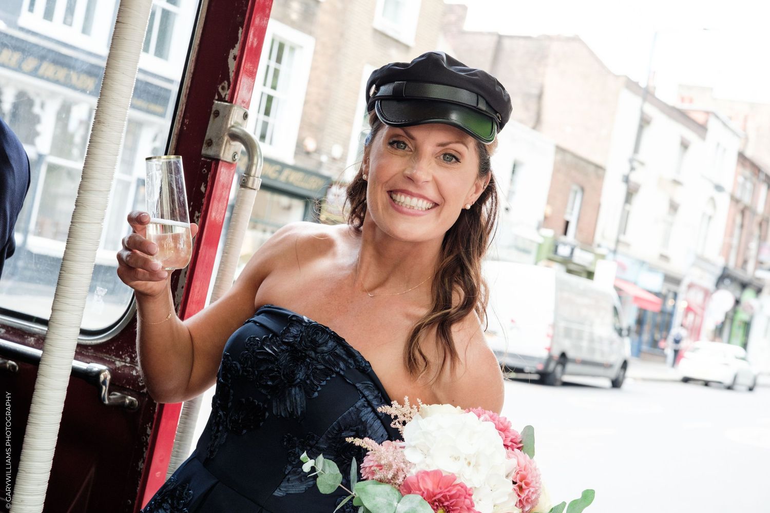 Smiling bride in navy strapless gown and black cap holding bouquet of pink and white flowers on a vintage bus.