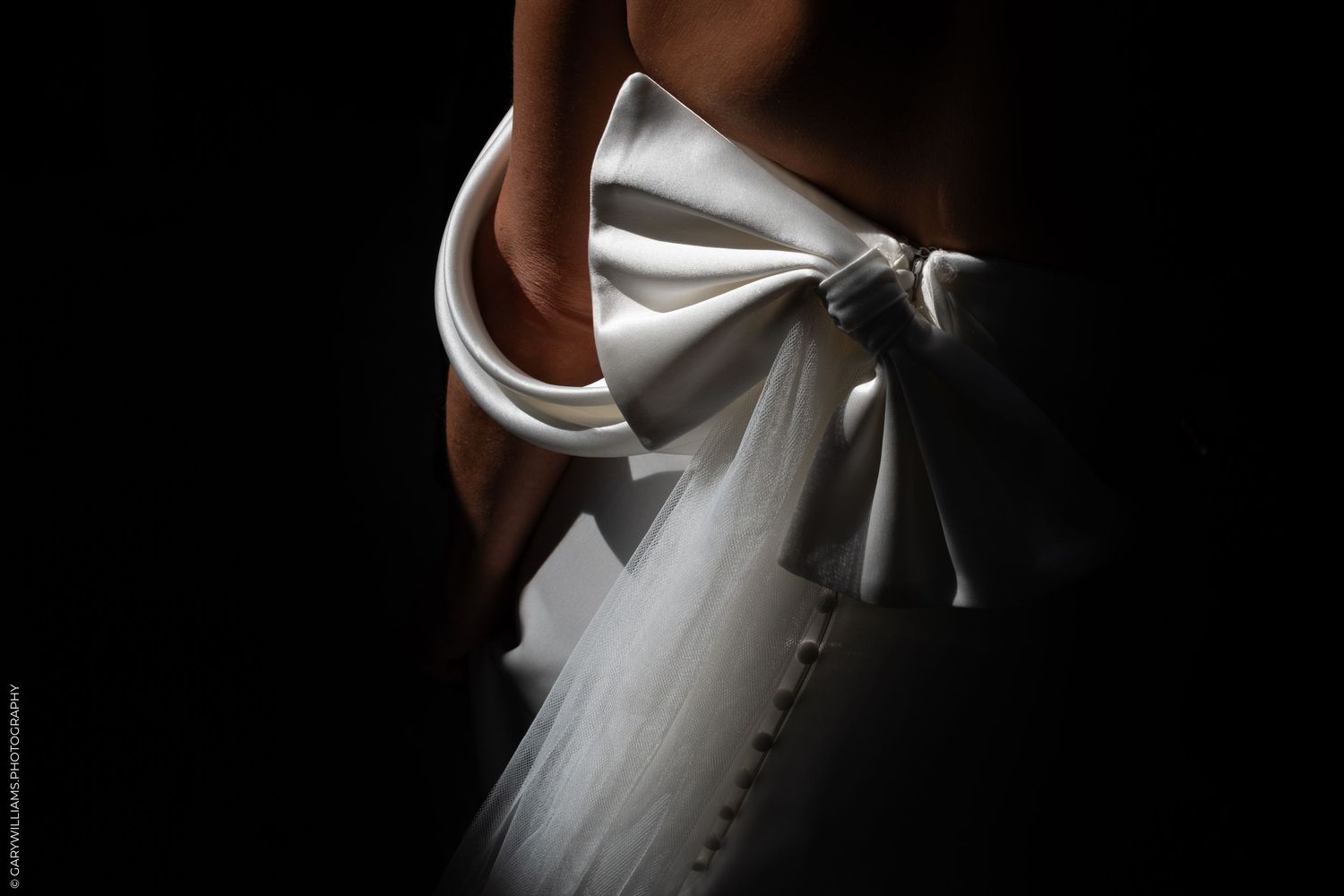 Close-up of an elegant white satin bridal gown bow detail against a dark background.